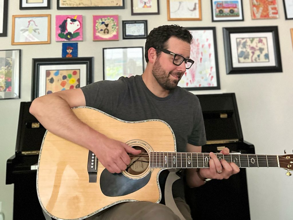 Bradley Kerr sitting with an acoustic guitar practicing in front of a piano and a gallery wall of art. 