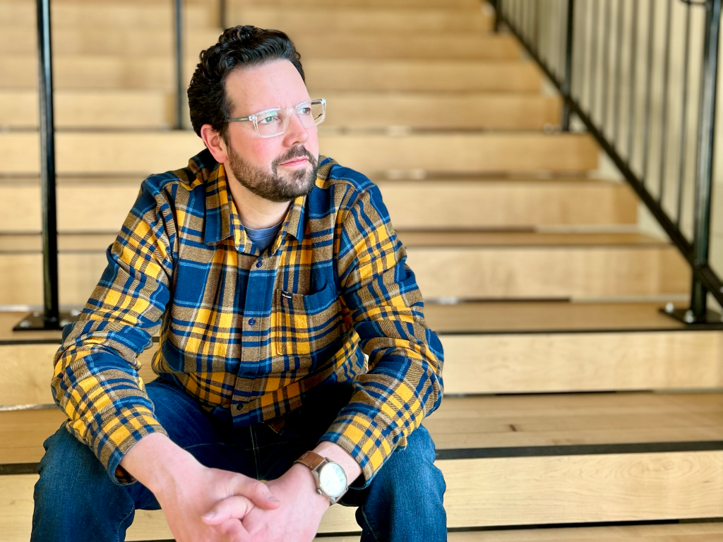 Headshot image of Bradley Kerr, in a yellow and blue plaid shirt, sitting on some steps at a music venue. 