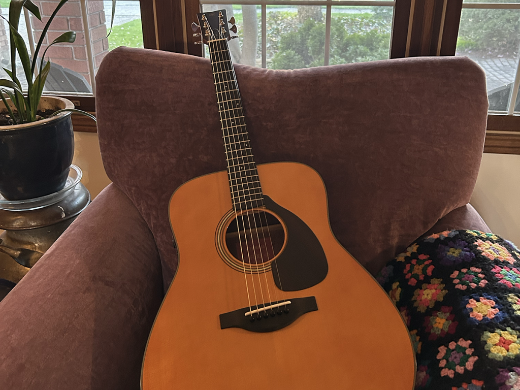 An acoustic guitar rests on a purple chair in a living room, with an old crocheted blanket beside it. 
